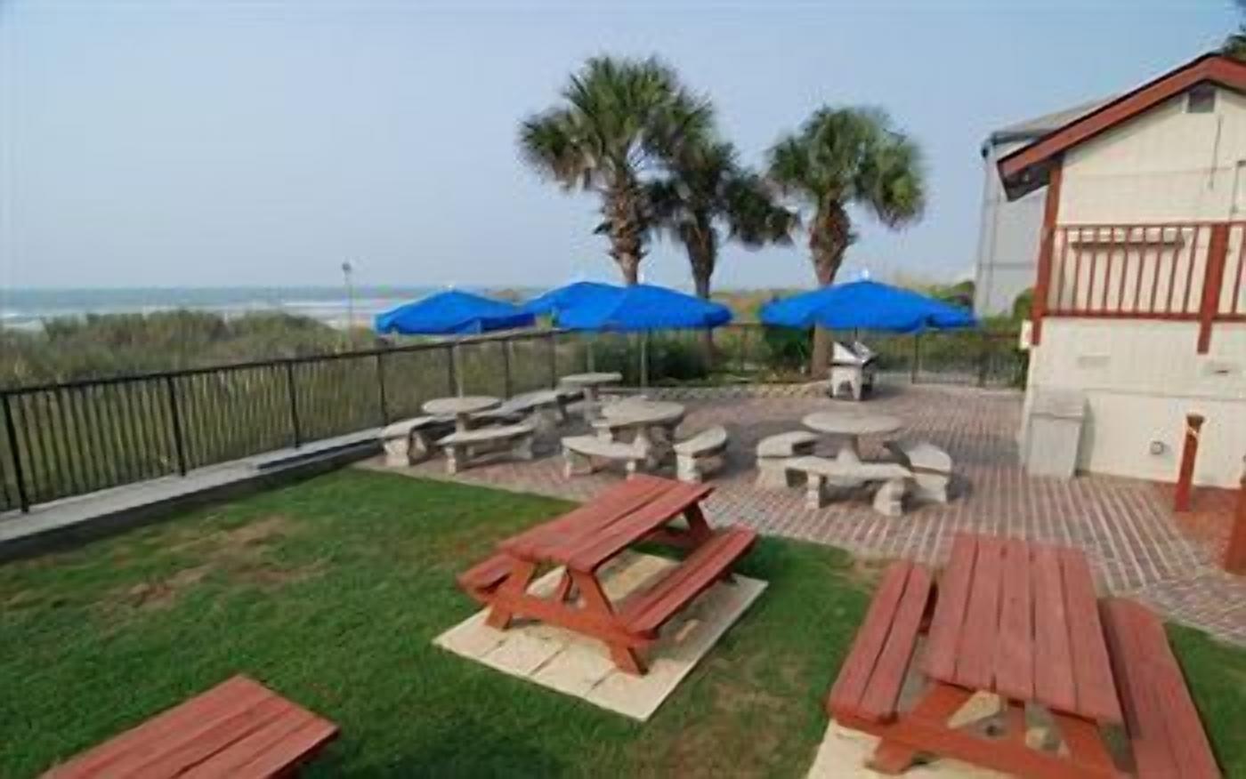 Eine Terrasse mit Picknicktischen und Sonnenschirmen, eine Rasenfläche und Blick auf den Strand.