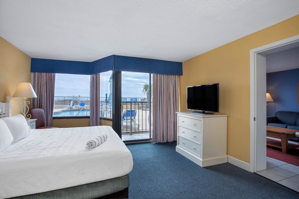 Een hotelkamer met een bed, een televisie, een bureau en uitzicht op een zwembad en palmbomen.