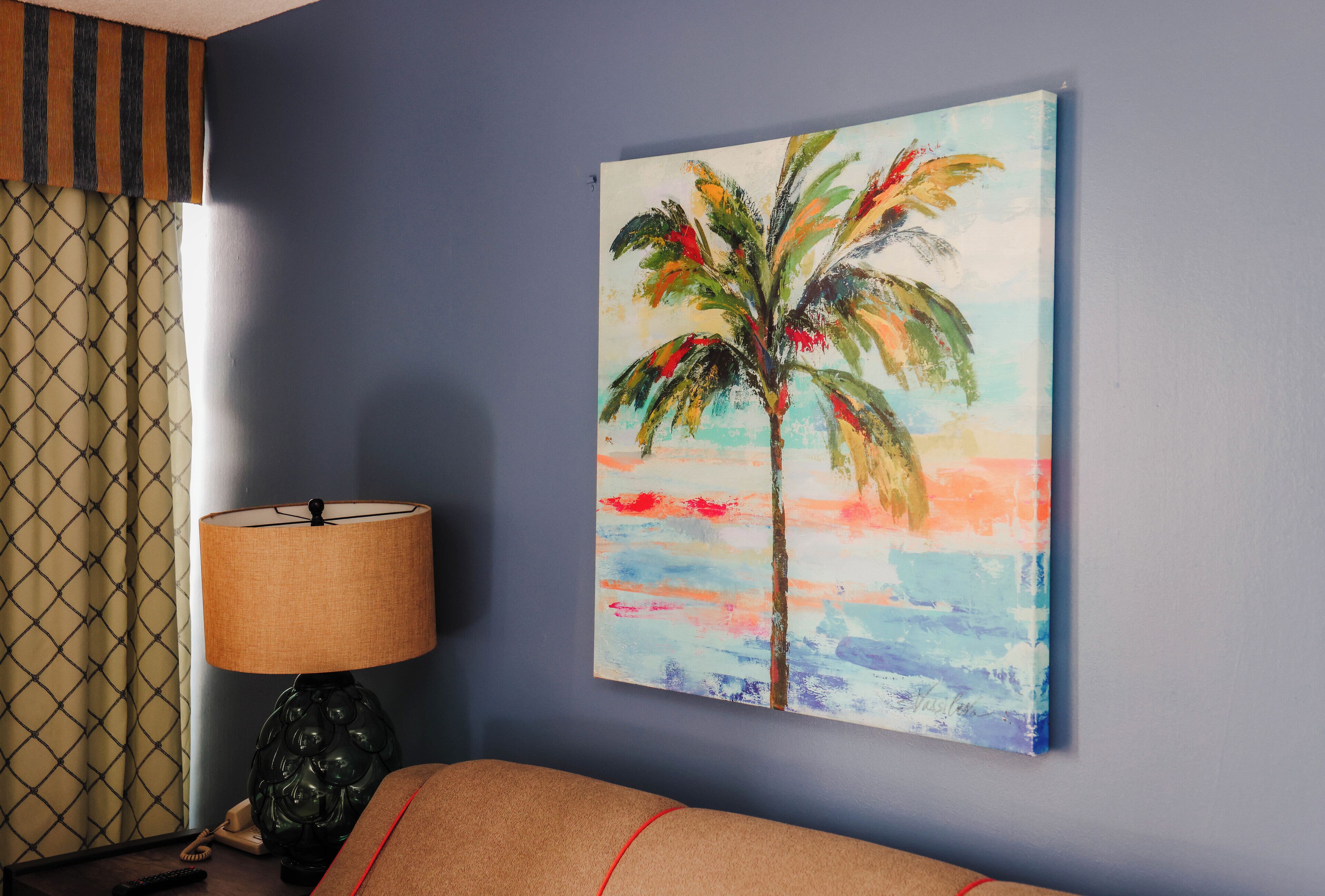 Quarto com parede azul, pintura de uma palmeira, luminária e cortina estampada.