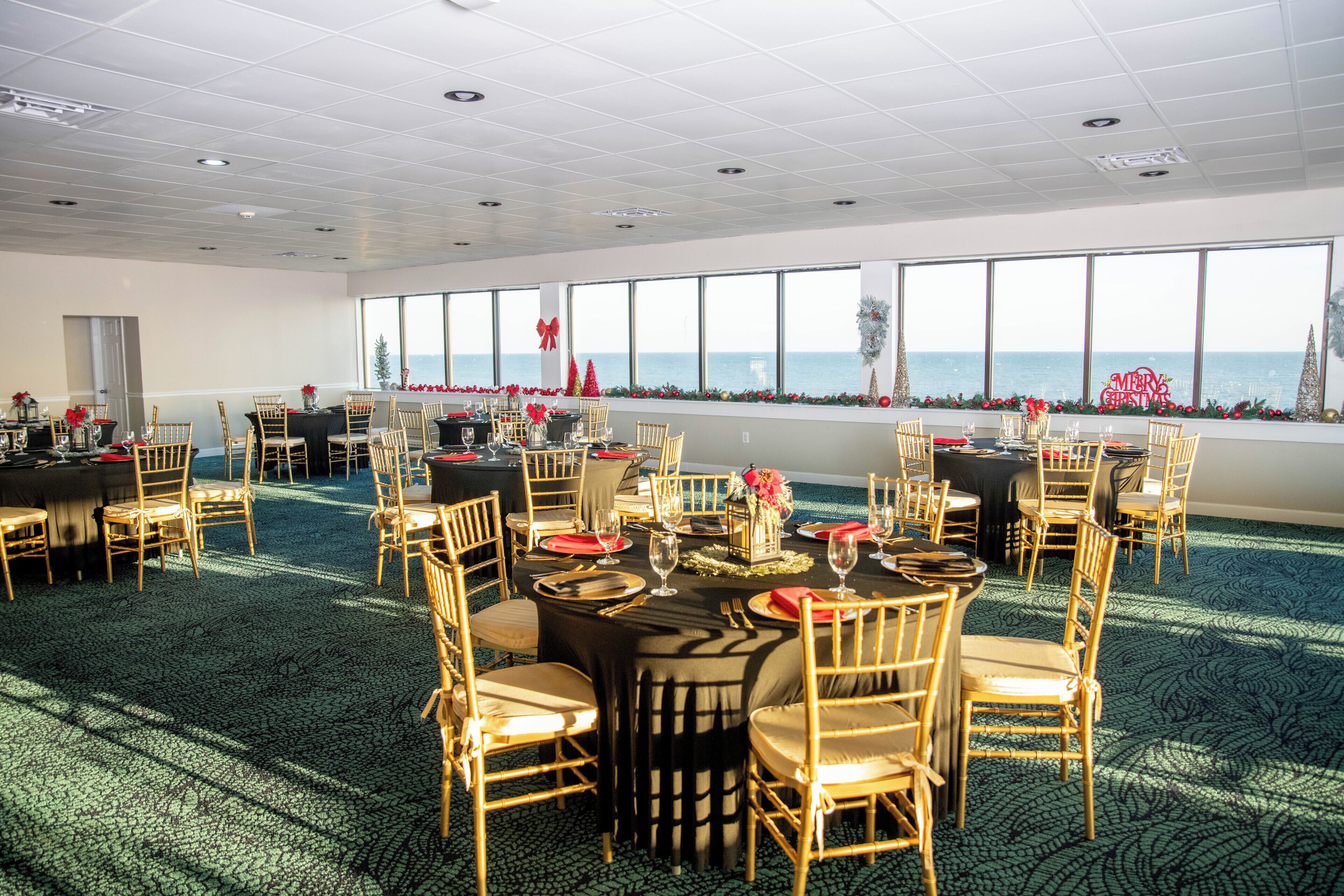 A large, well-lit room with multiple tables set for dining, large windows with a view of the sea, and a carpeted floor.