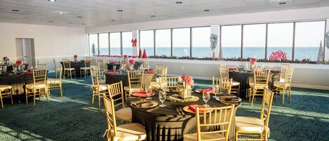 A large, well-lit room with multiple tables set for dining, large windows with a view of the sea, and a carpeted floor.