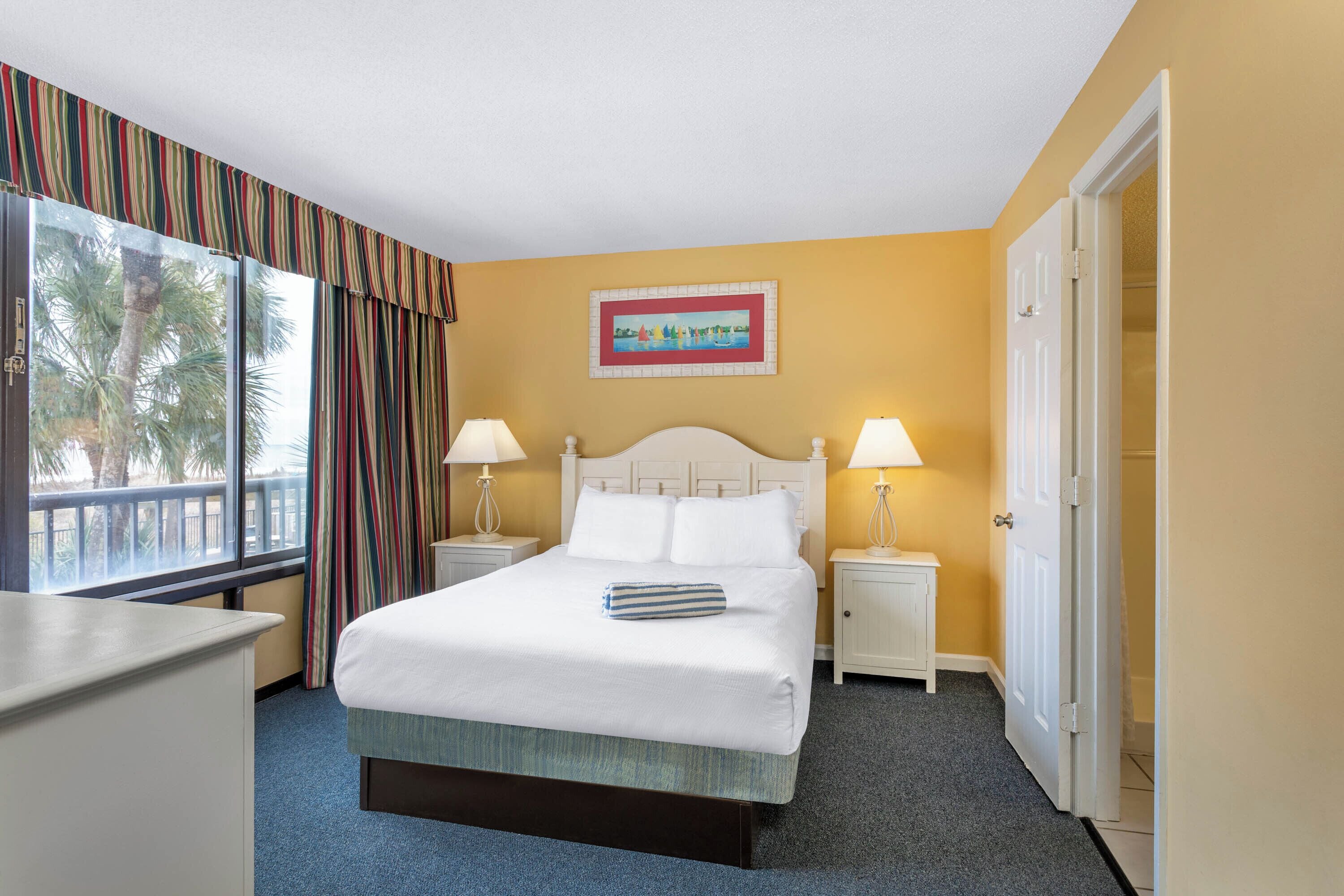A hotel room with a bed, two bedside tables with lamps, a window with curtains, a painting on the wall, and a door.