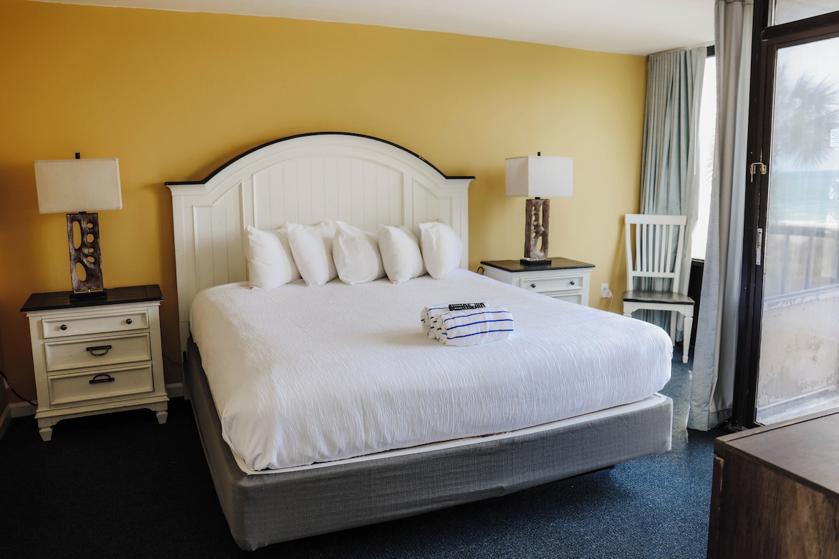 Quarto de hotel com uma cama grande, duas mesas de cabeceira com abajures, uma cadeira e uma janela com cortinas.