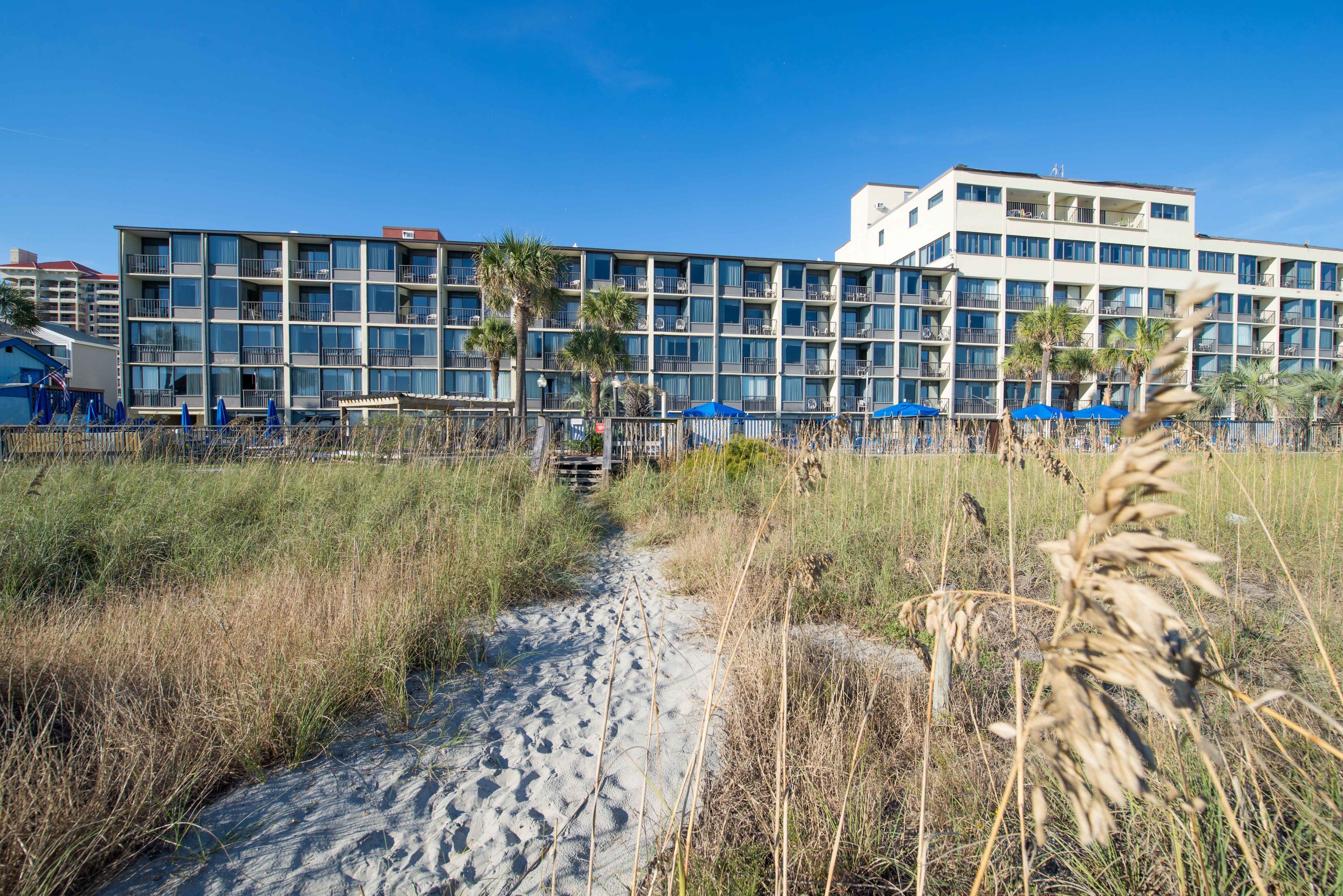 Un hotel frente a la playa con varios balcones, un sendero de arena y palmeras.
