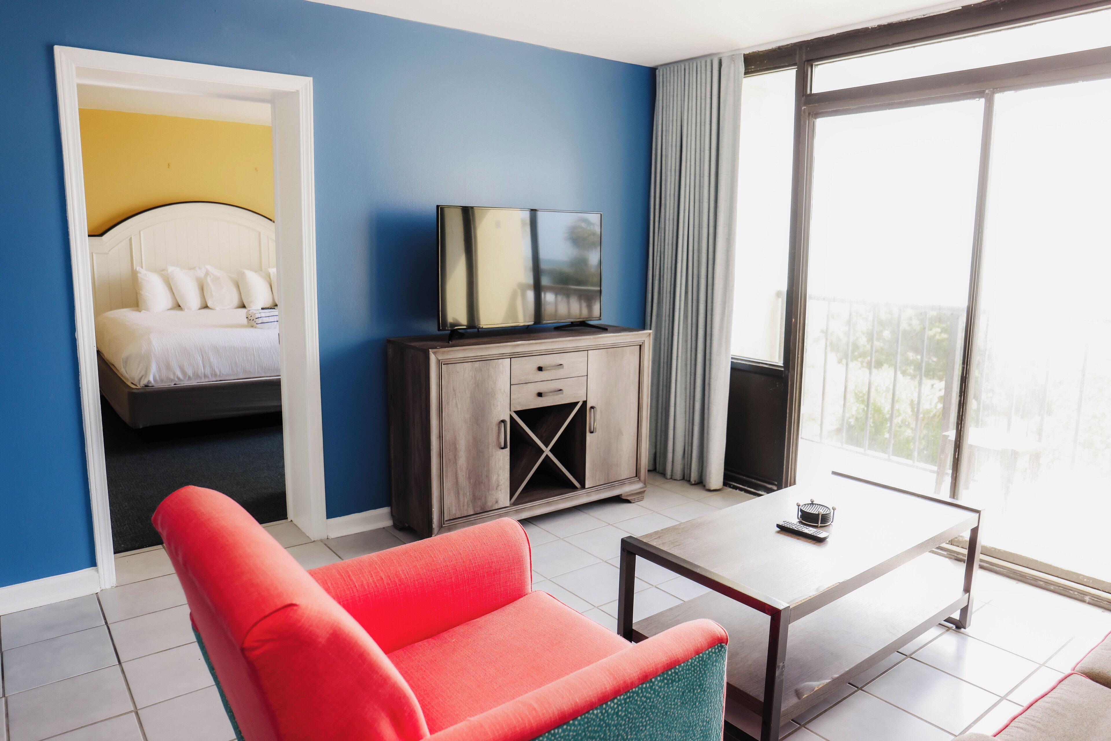 A hotel room with a bed, a television, a red chair, a small table, and a view of the outdoors.