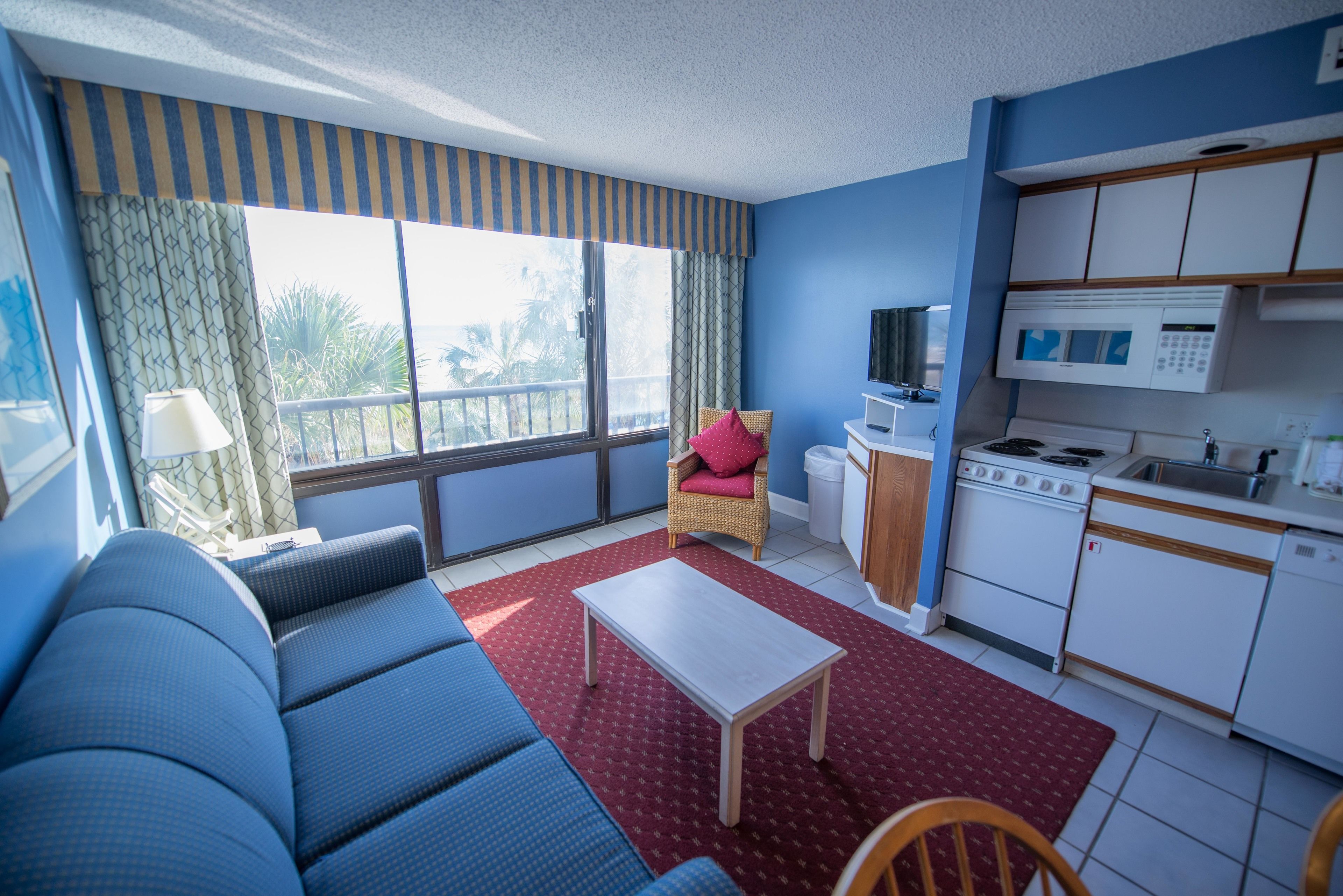 A room with a sofa, a small table, a kitchen area with a microwave, and a window with curtains.