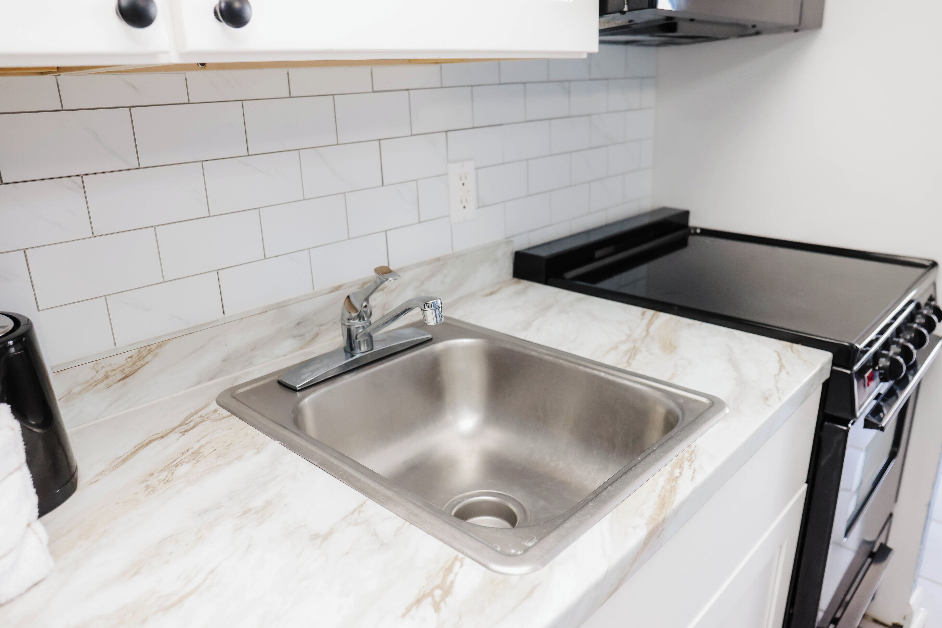 Una cocina con un lavamanos de acero inoxidable, una hornilla negra y un salpicadero de azulejos blancos.