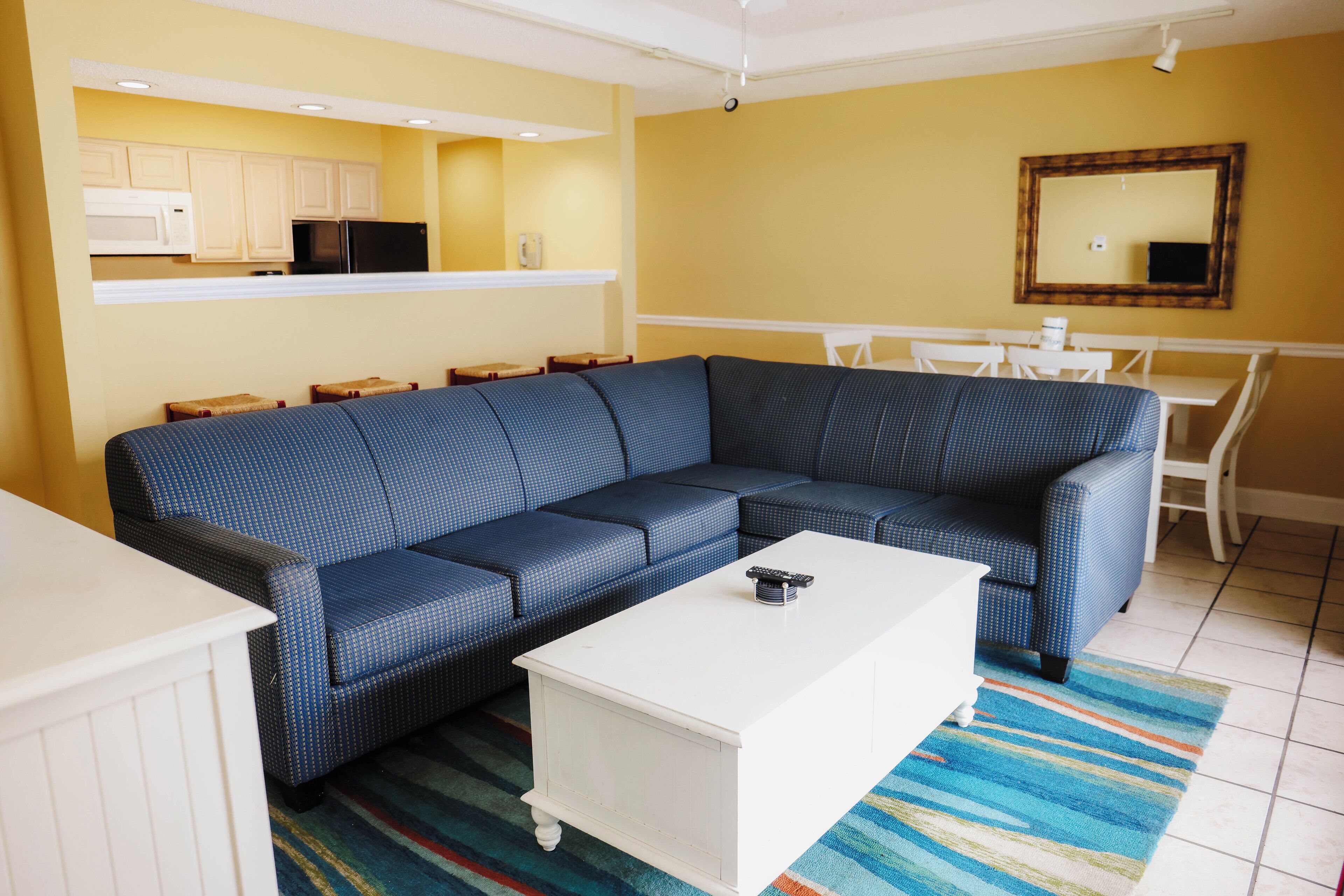 A living room with a blue sofa, white coffee table, and a kitchen area with white cabinets and appliances.