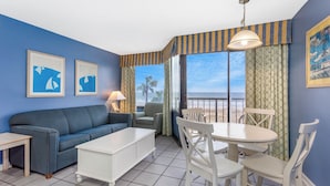 A room with a blue wall, a sofa, a dining table with chairs, a lamp, and a view of the beach.