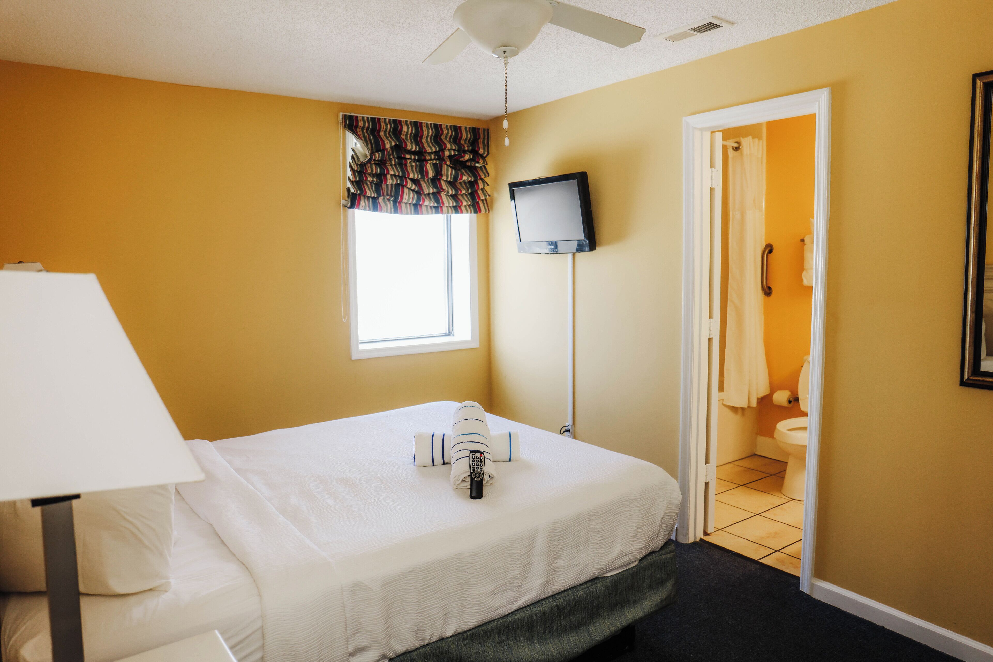 Una habitación de hotel con cama, televisor montado en la pared, ventana con cortinas y puerta que da al baño.