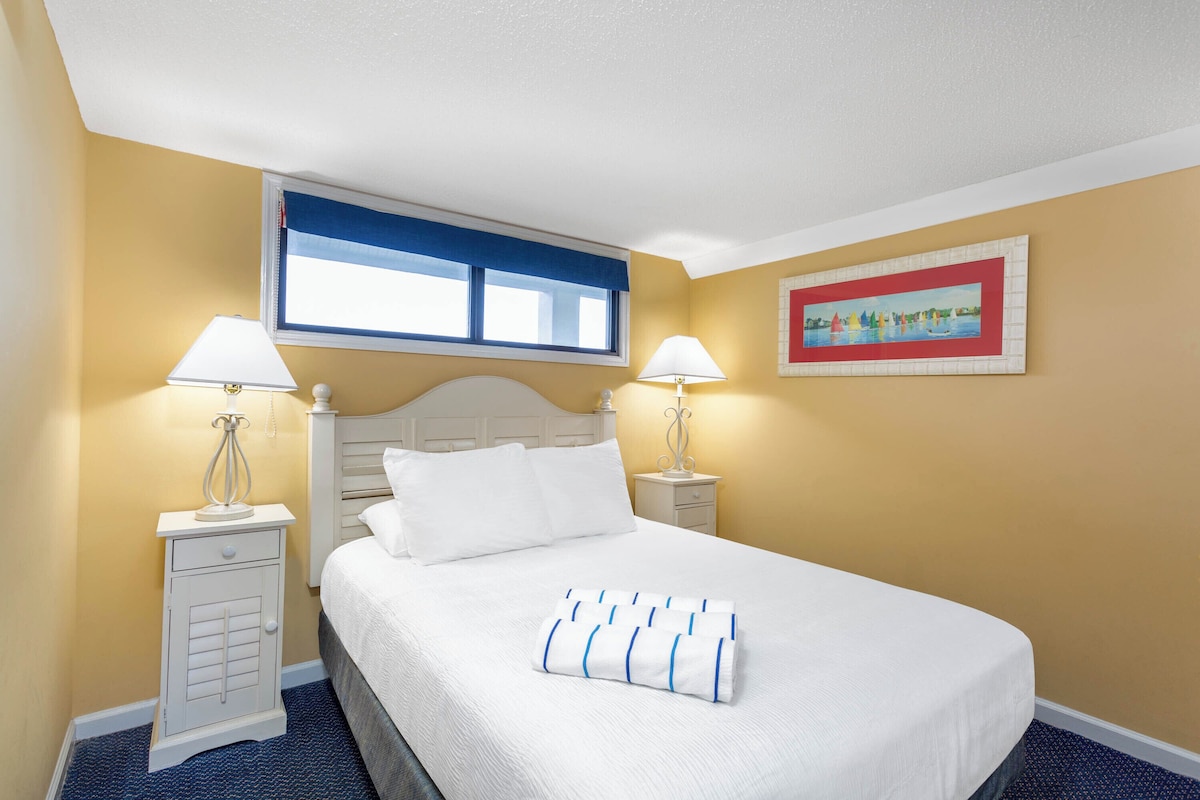 Quarto de hotel com cama, duas mesas de cabeceira, luminárias, janela com cortinas azuis e um quadro na parede.