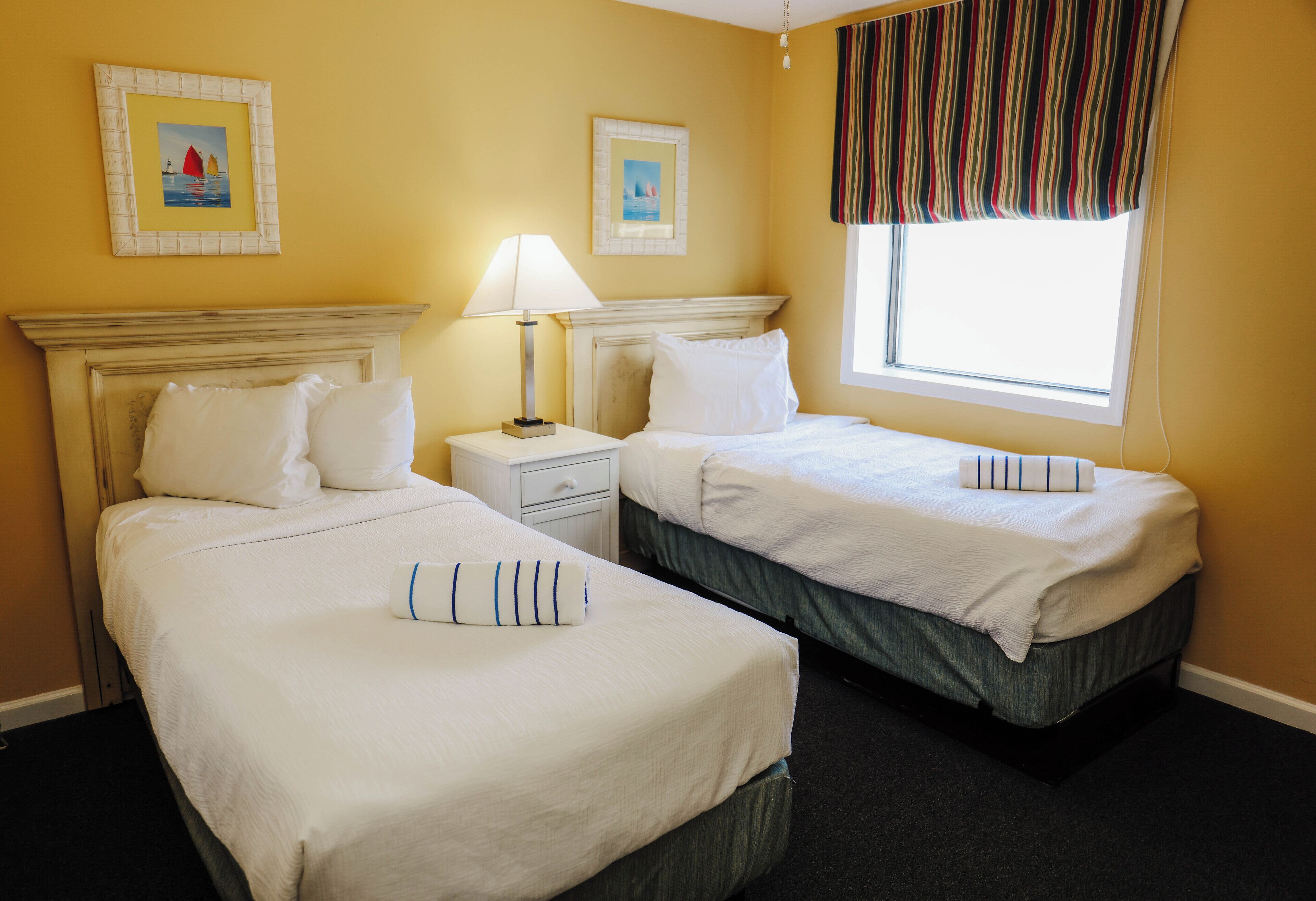 A hotel room with two beds, a nightstand, a lamp, and a window with curtains.