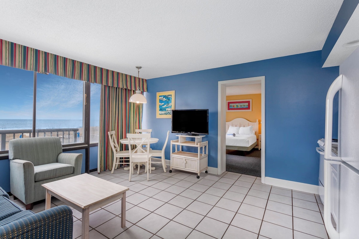 Quarto de hotel com uma grande janela, televisão, cama, escrivaninha e uma cadeira.