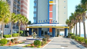L’entrée d’un hôtel, ornée d’une enseigne portant le nom « ATLANTICA », est entourée de palmiers et d’un jardin bien entretenu.