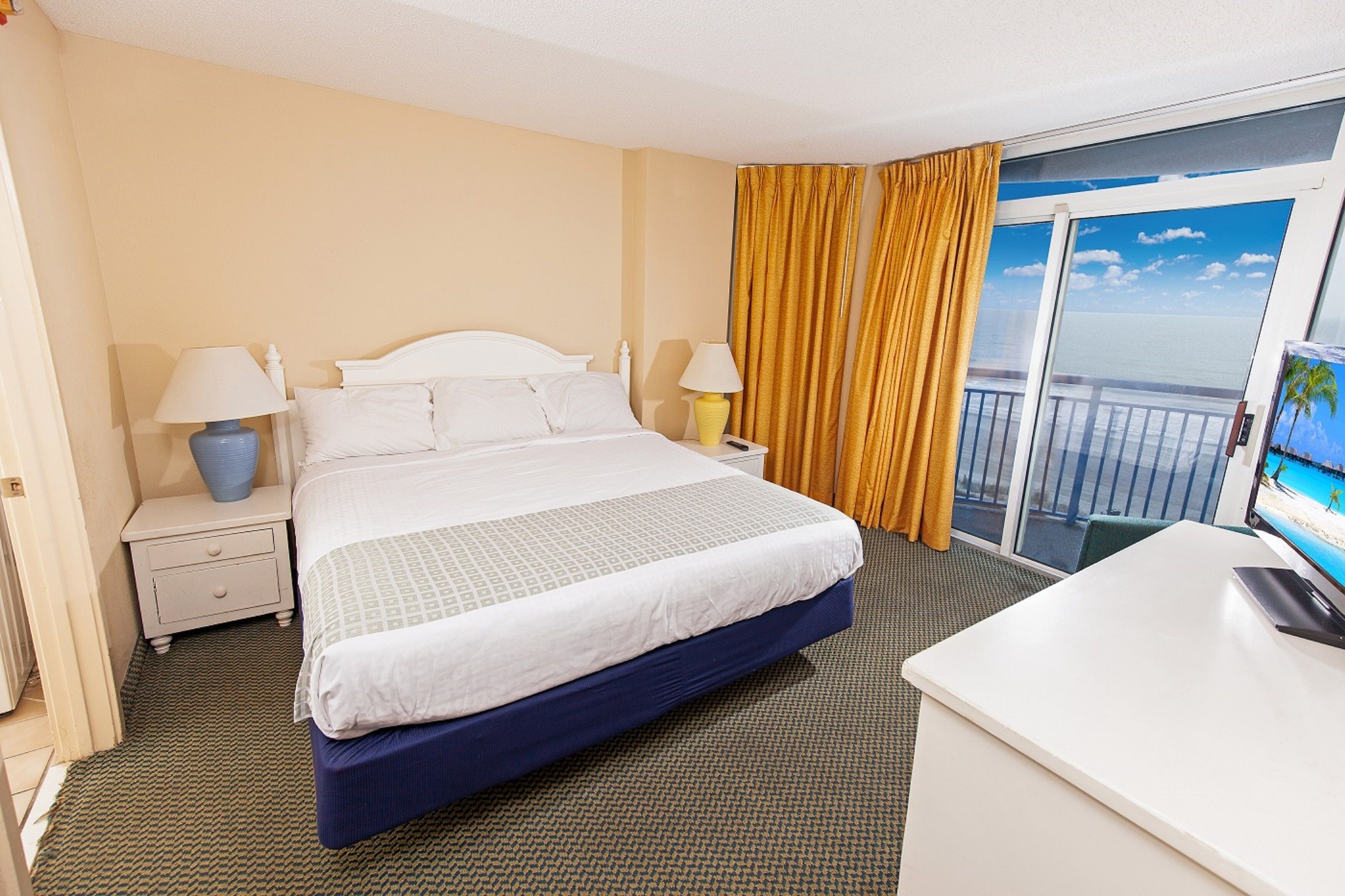 A hotel room with a bed, bedside tables, a desk, and a view of the ocean through a balcony.