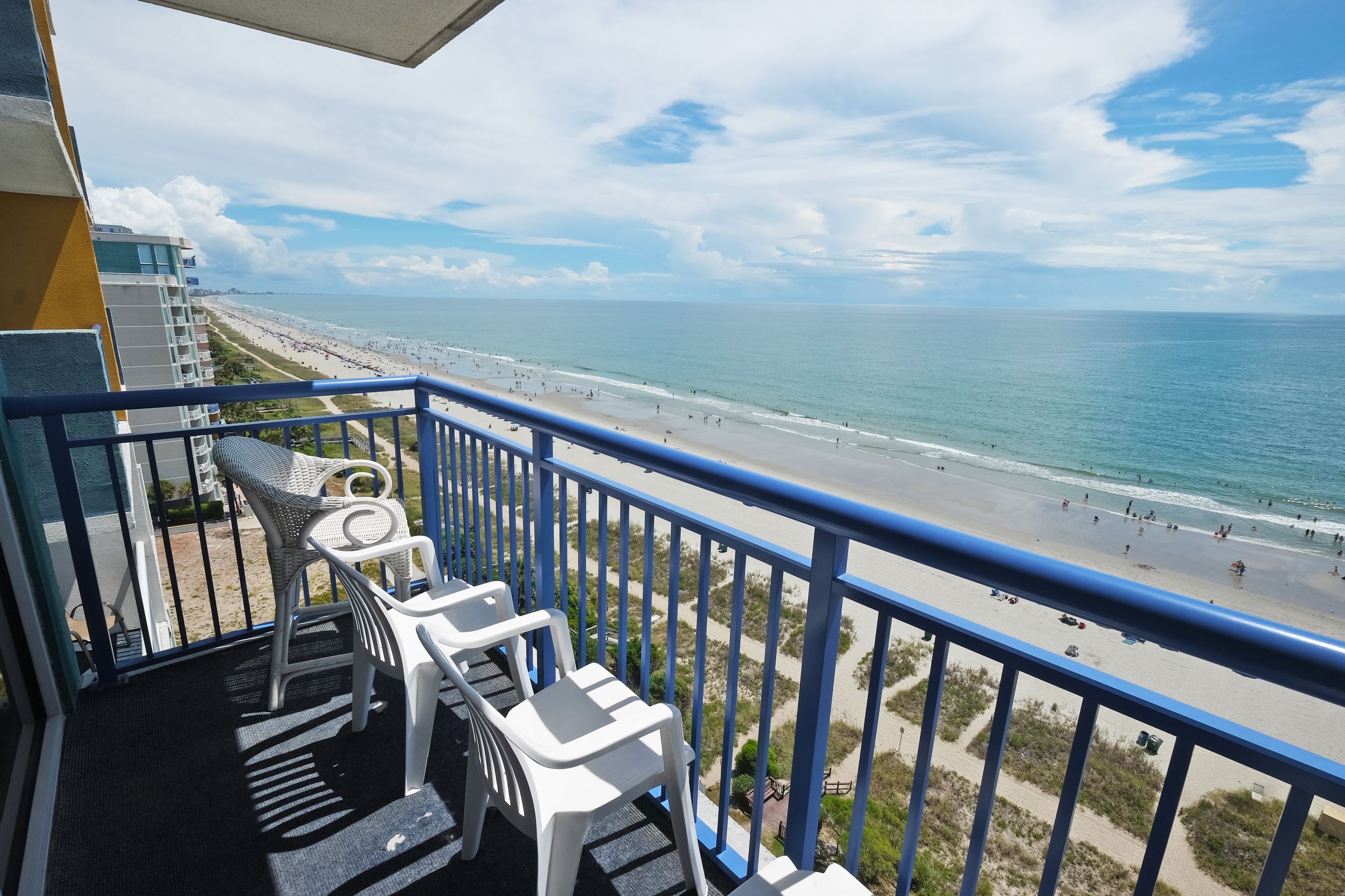 Un balcone con sedie in plastica bianca, una ringhiera blu e vista su una spiaggia con persone e cielo sereno.