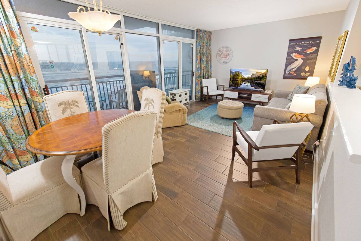 Uma sala de estar ampla, com uma mesa de jantar de madeira, assentos confortáveis, televisão e vista para o oceano através de grandes portas deslizantes.