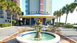 L’entrée d’un hôtel, avec une fontaine, des palmiers et une enseigne indiquant « ATLANTICA ».