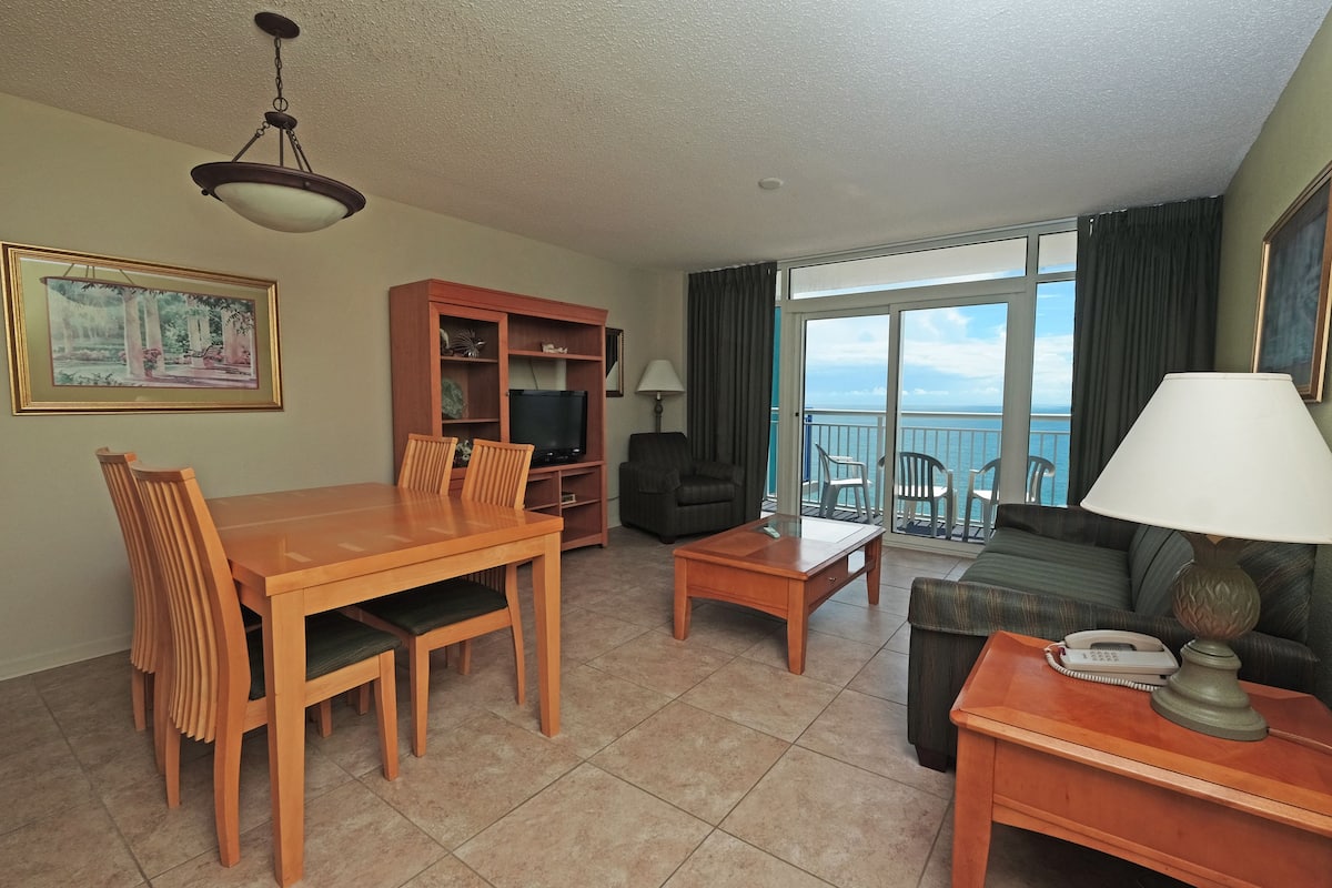 Quarto de hotel com mesa de jantar, cadeiras, sofá, mesinha de centro, televisão e varanda com vista para o mar.