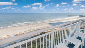 Un balcon avec des balustrades blanches donnant sur une plage, avec une jetée visible au loin.