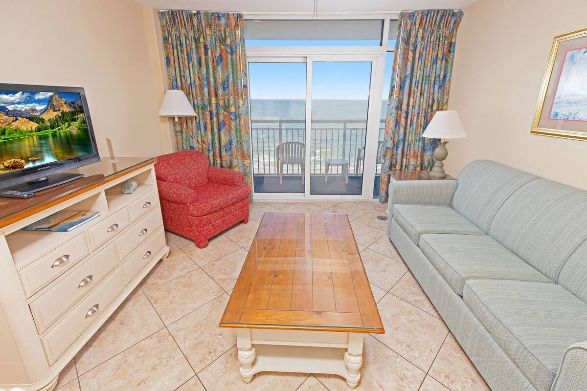 Uma sala de estar com televisão, uma poltrona vermelha, um sofá cinza, uma mesa de centro de madeira e uma varanda com cortinas.