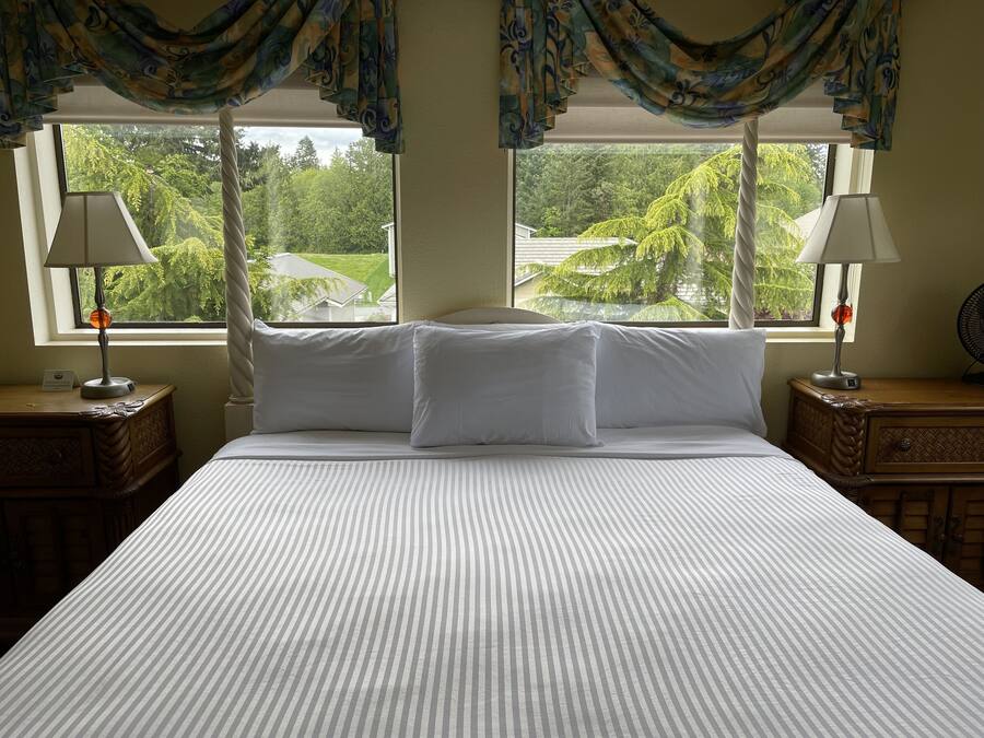 A bed with white linens, two bedside tables with lamps, and a view of greenery through large windows.