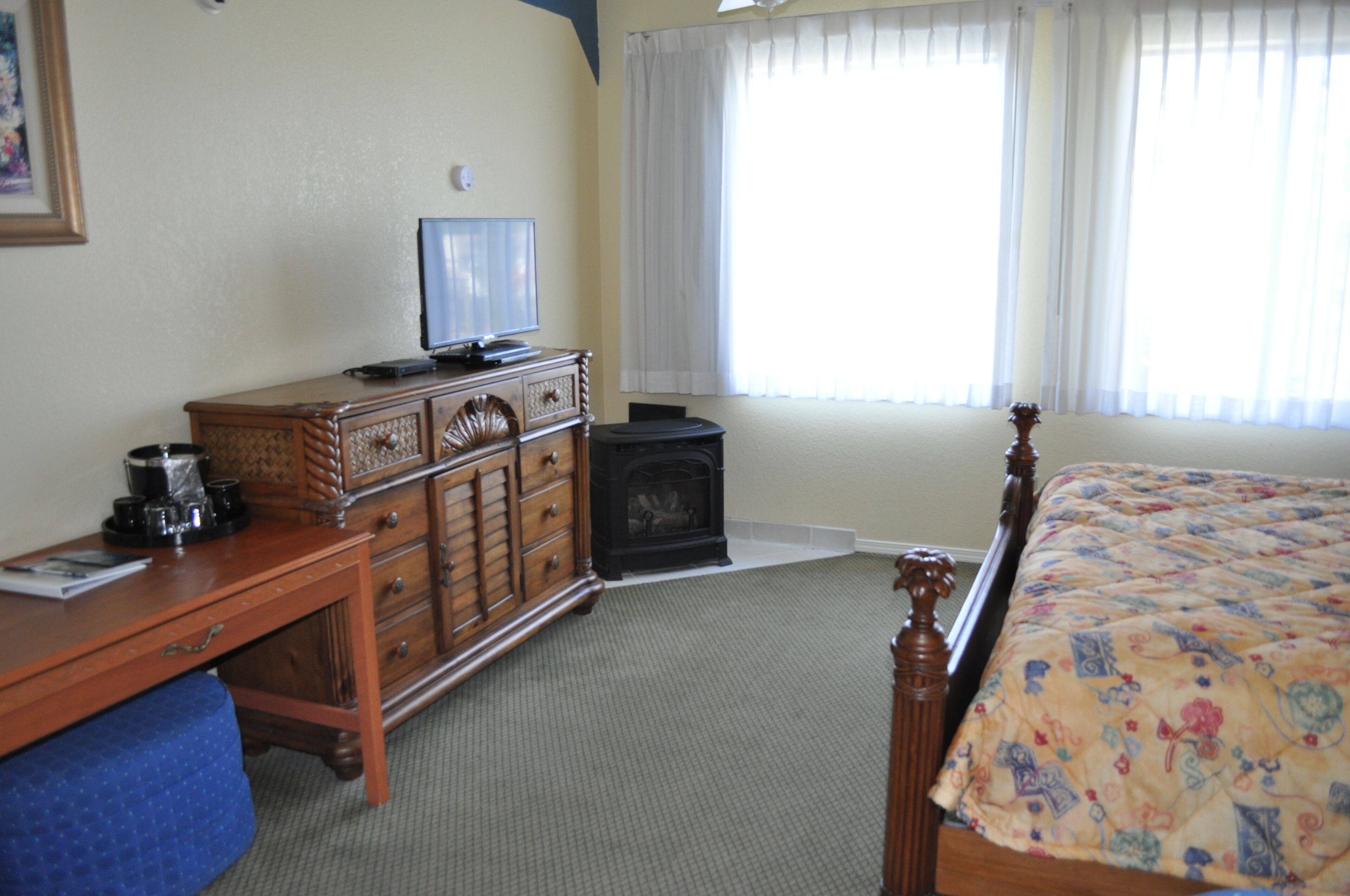 A hotel room with a wooden dresser, a television, a small black heater, a bed with a floral bedspread, and a window with white curtains.