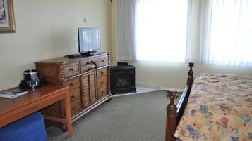 A hotel room with a wooden dresser, a television, a small black heater, a bed with a floral bedspread, and a window with white curtains.