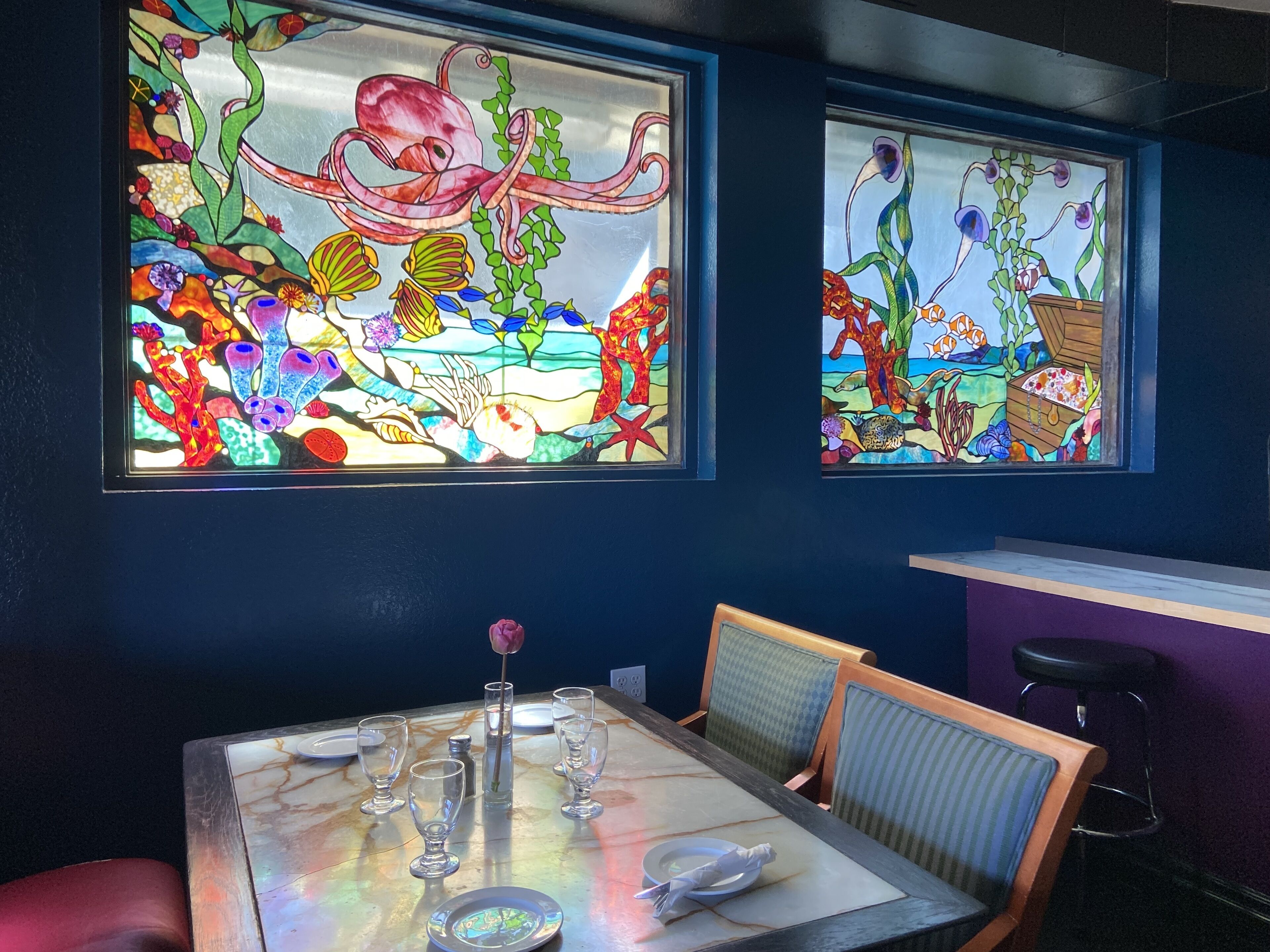 A dining area with a table set for two, a vase with a single flower, and two large stained glass windows depicting underwater scenes.