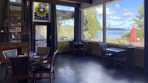 A dining area with wooden furniture, a large window with a view of a lake and boats, and an exit sign. - Old Alcohol Plant Hotel (Port Hadlock)