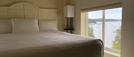 A large bed with white linens, a bedside lamp, and a window with a view of boats on a body of water.