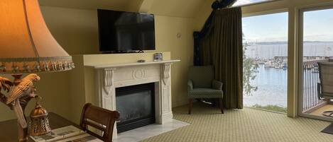 A room with a fireplace, a television, a dining table with chairs, and a view of a marina through large windows.