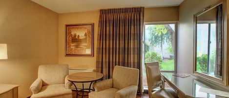 A room with a wooden desk, two armchairs, a small round table, and a large window with curtains.