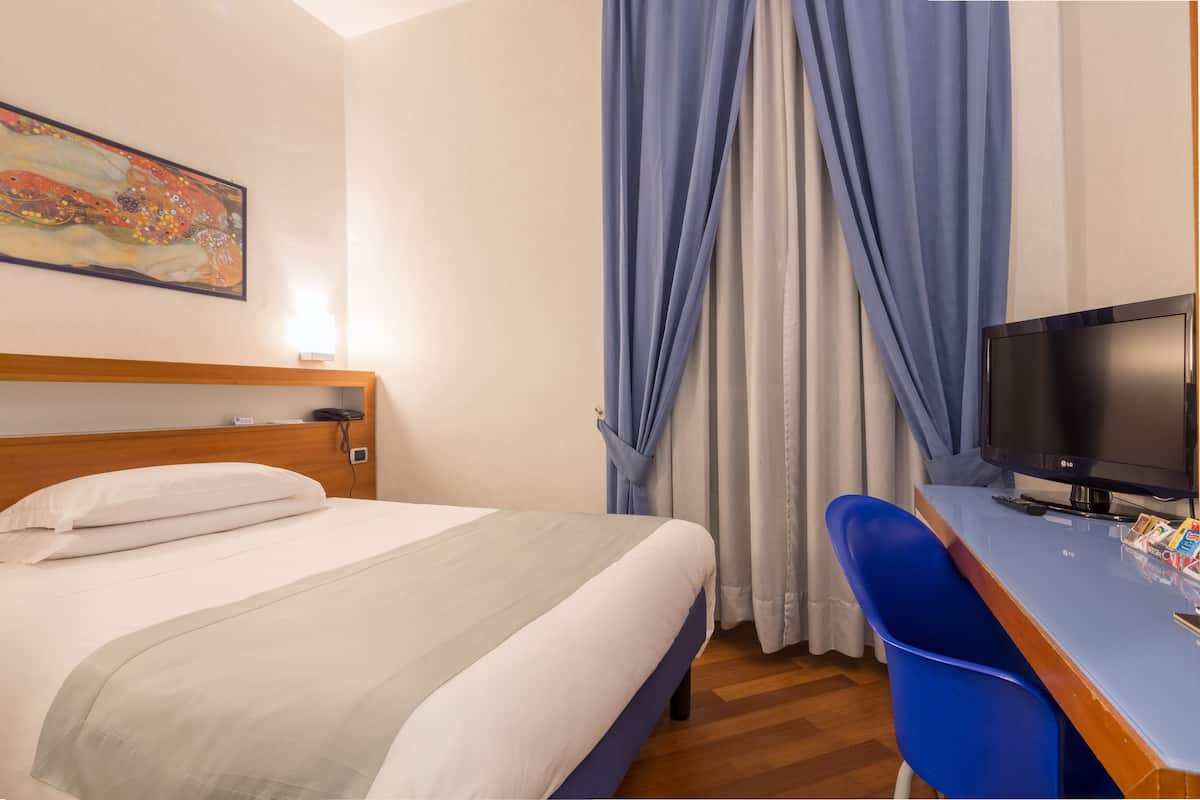 Habitación de hotel con cama, escritorio con televisor, un cuadro en la pared y ventana con cortinas.