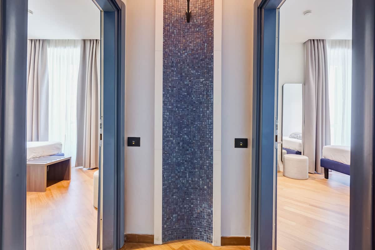 Una habitación de hotel moderna con suelo de madera, un ventanal con cortinas, una cama, una mesita de noche y una pared con textura.