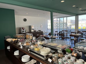 Buffet de café da manhã com diversos alimentos, uma máquina de café e um relógio na parede.