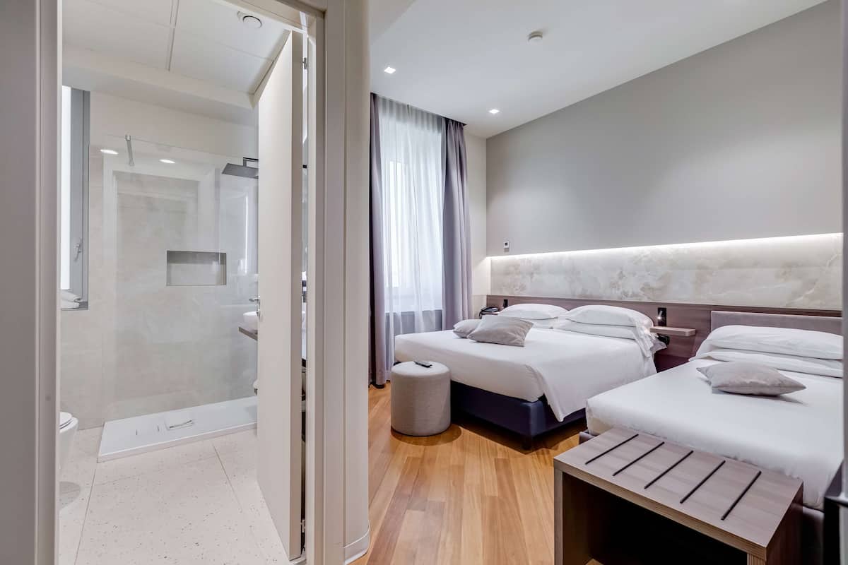 Una habitación de hotel moderna con cabina de ducha de vidrio, suelo de madera, dos camas con ropa blanca, una mesita de noche y un pequeño taburete.