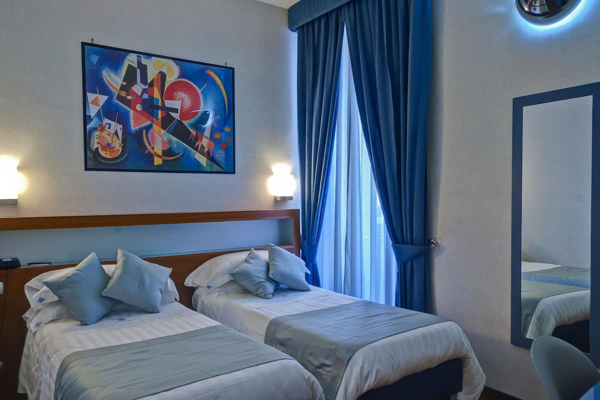 Habitación de hotel con dos camas, un cuadro en la pared, un ventanal grande con cortinas azules y un espejo.