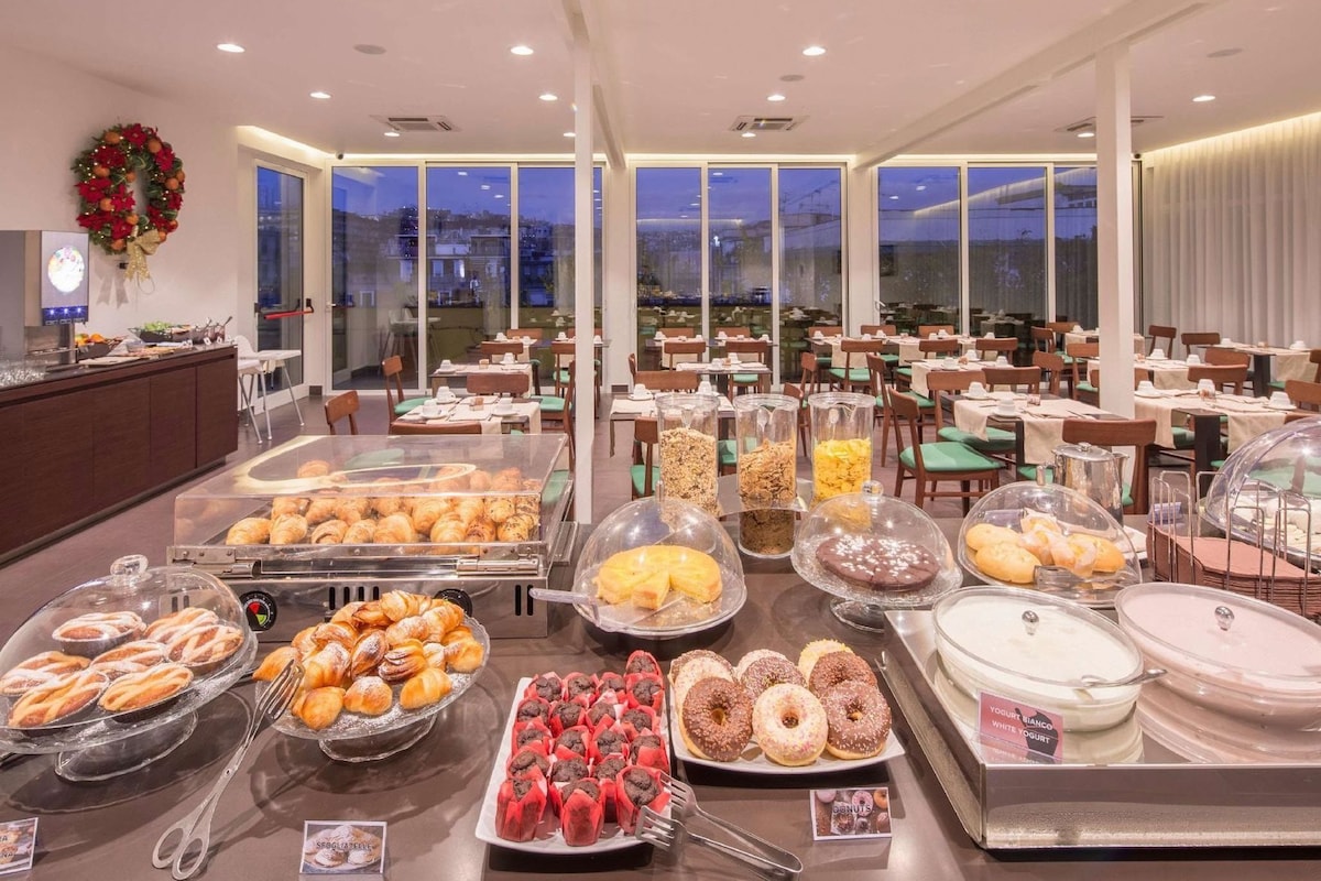 A buffet with various food items, including pastries, donuts, and yogurt, in a well-lit dining area with large windows.