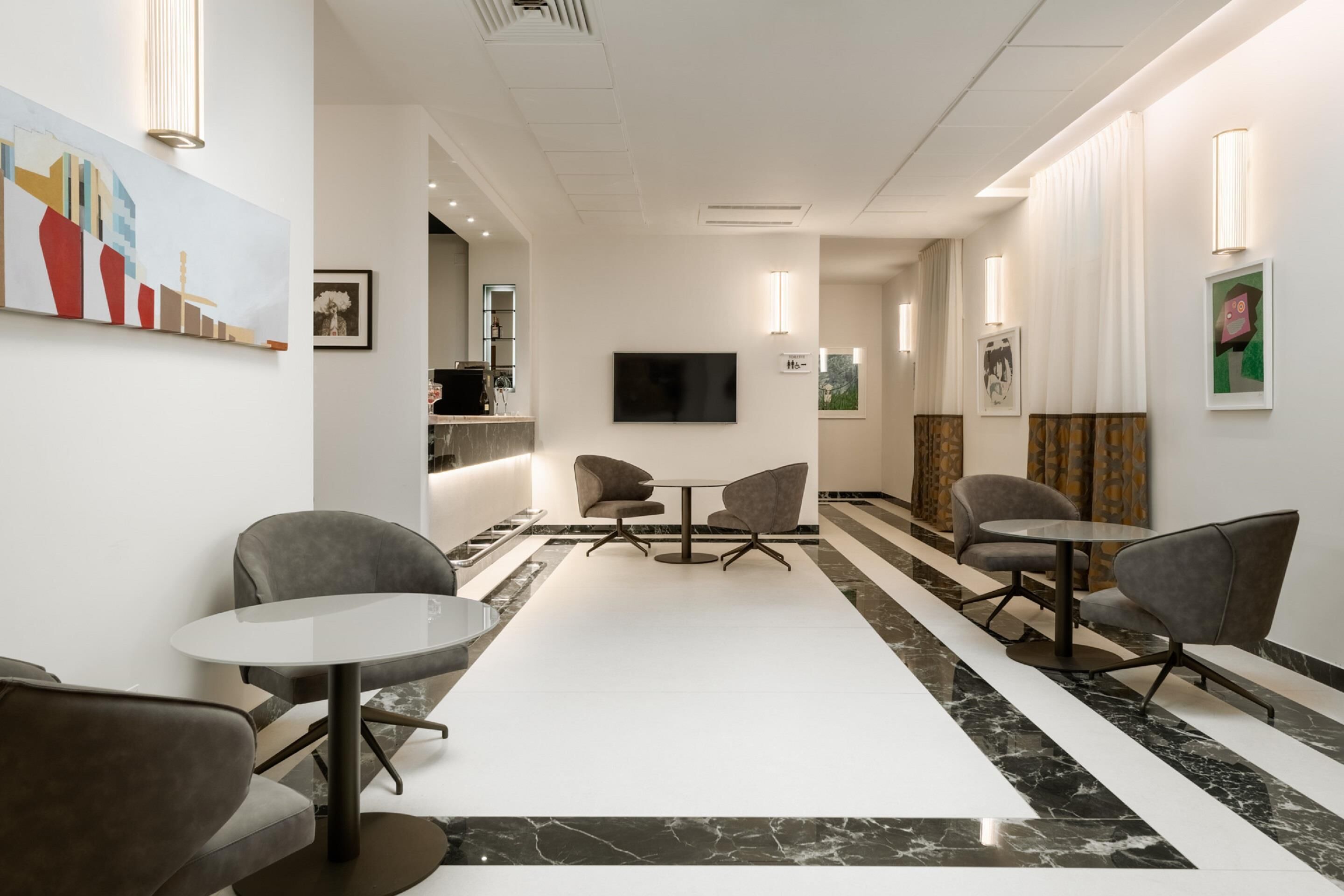 A modern hotel lobby with a marble floor, grey upholstered chairs, a flat-screen TV, and artwork on the walls.