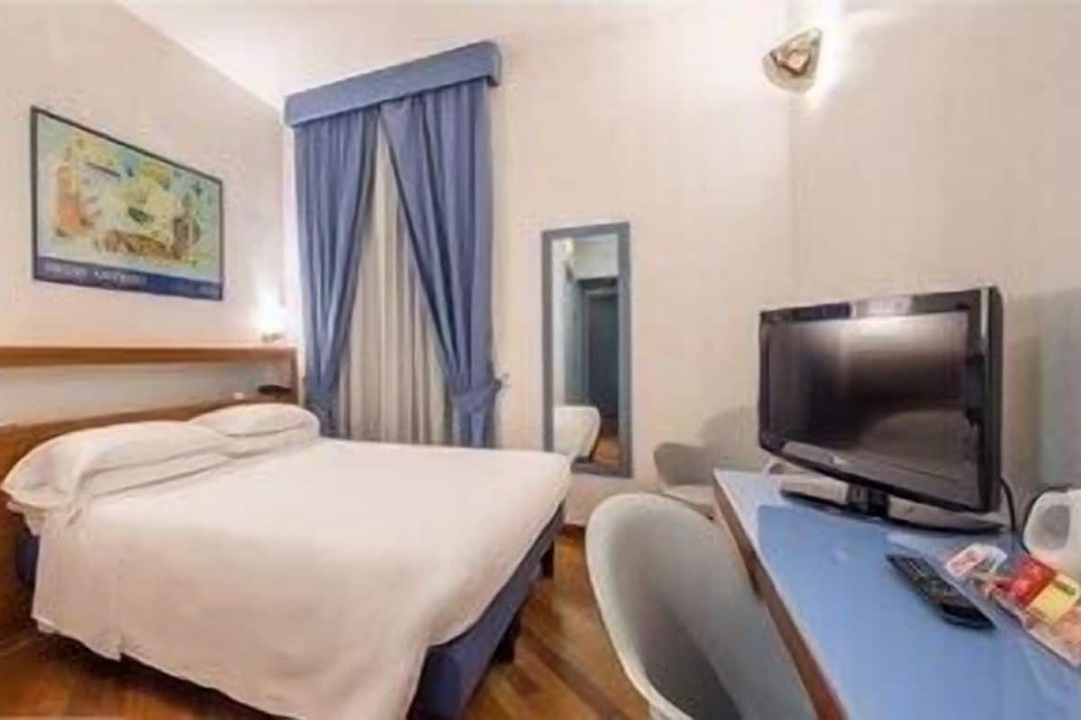Una habitación de hotel con cama, televisión, escritorio con silla y ventana con cortinas.