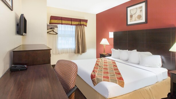 Habitación de hotel con cama, escritorio, silla, televisor, ventana con cortinas y lámpara.