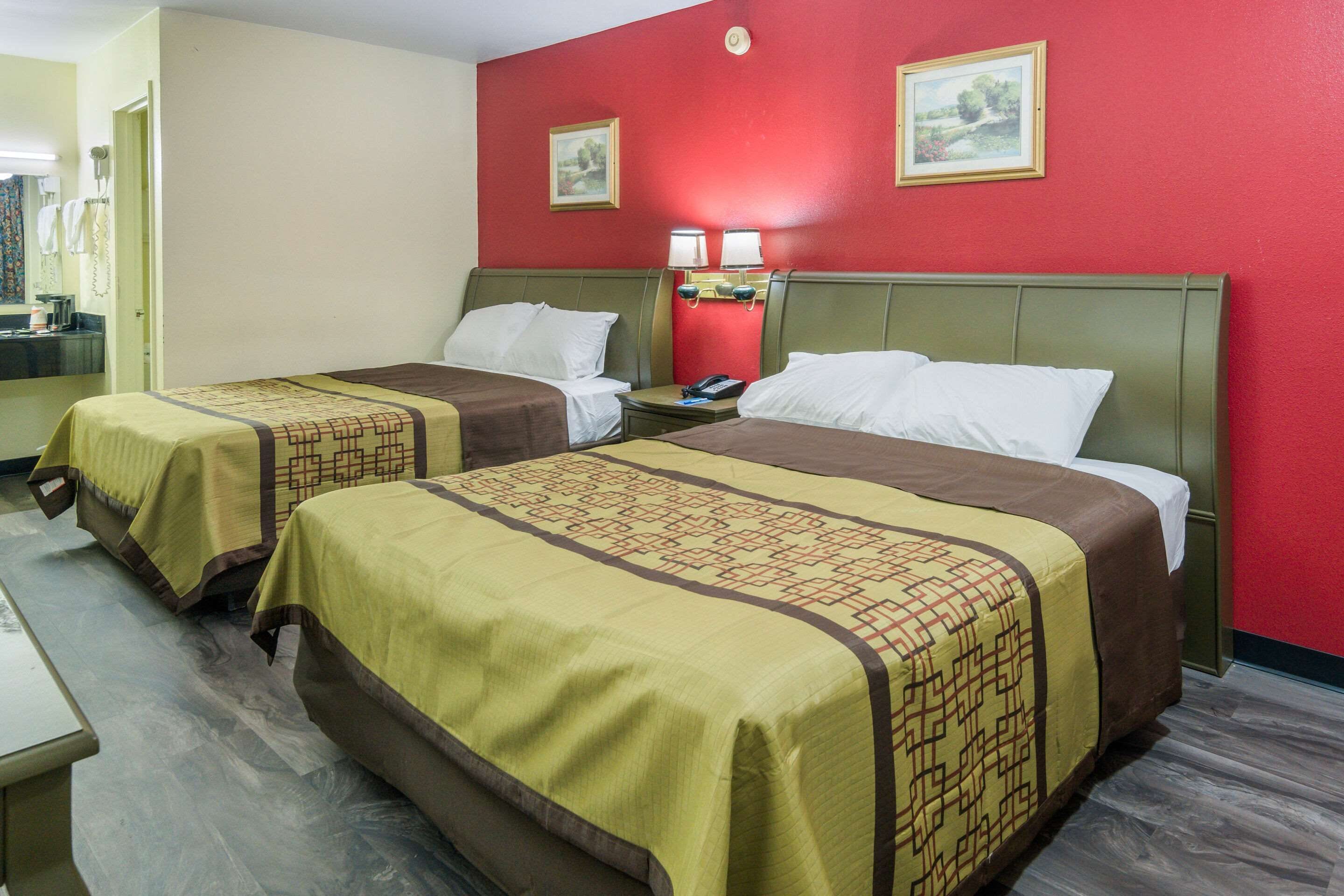A hotel room with two beds, each with a patterned bedspread, a nightstand with a lamp, and a red wall with two framed pictures.