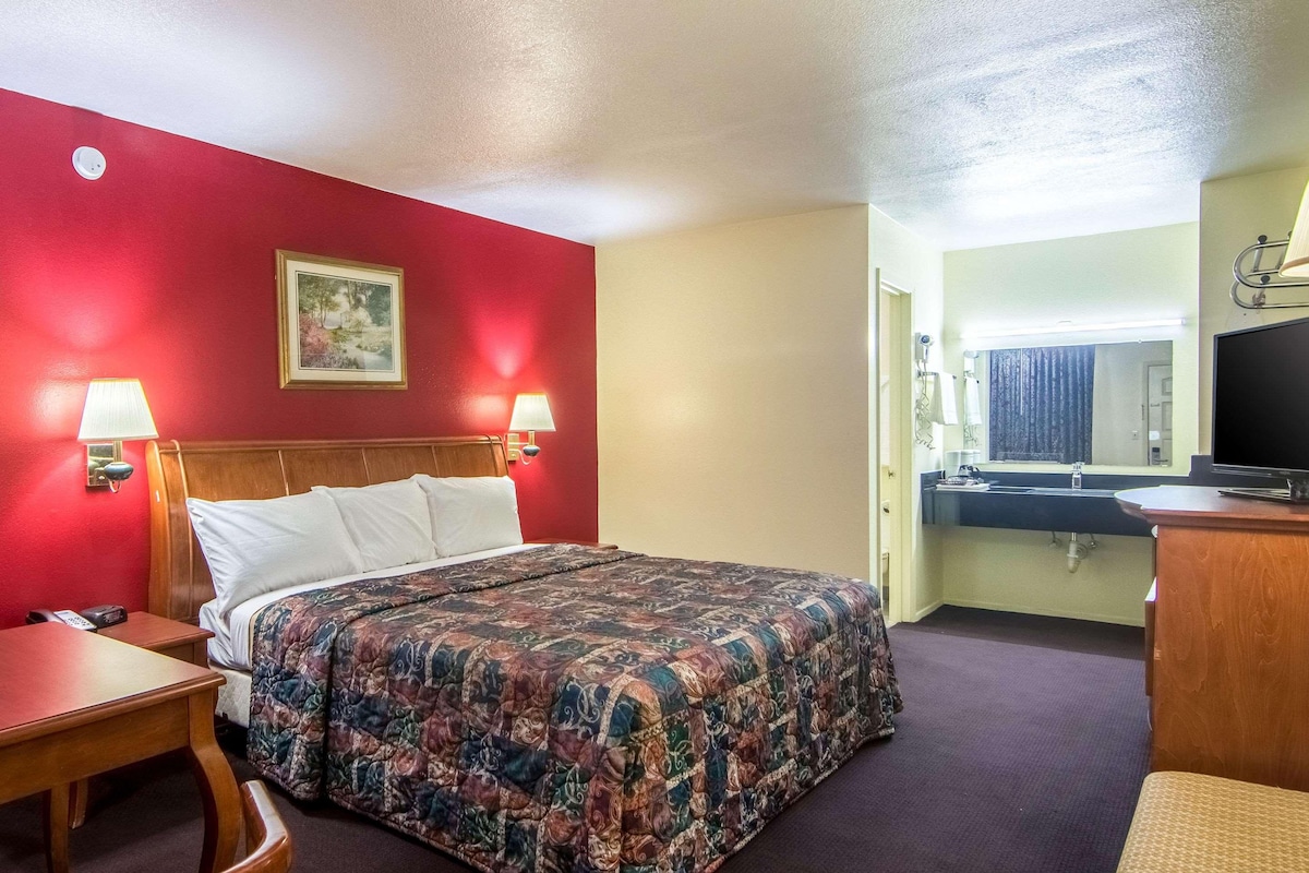 Habitación de hotel con cama, lámparas de noche, escritorio, silla, televisor, baño con lavamanos y espejo, y un cuadro en la pared.