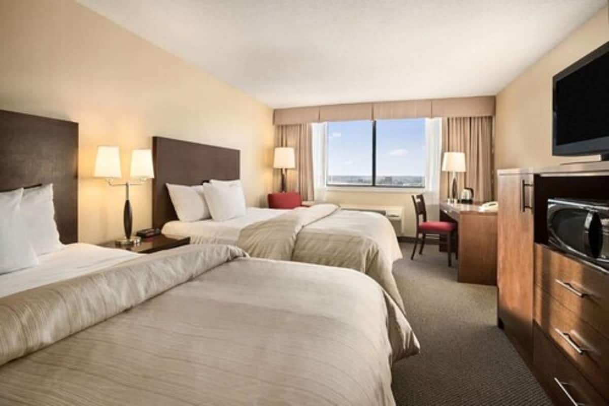 Quarto de hotel com duas camas, uma mesa, uma cadeira, uma televisão e janela com vista para a cidade.
