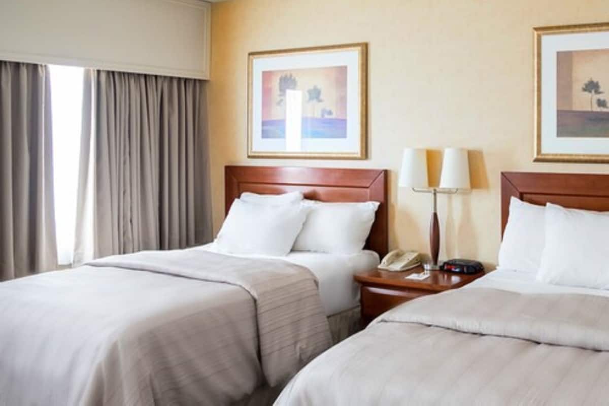 Quarto de hotel com duas camas, uma mesa de cabeceira com luminária e quadros nas paredes.