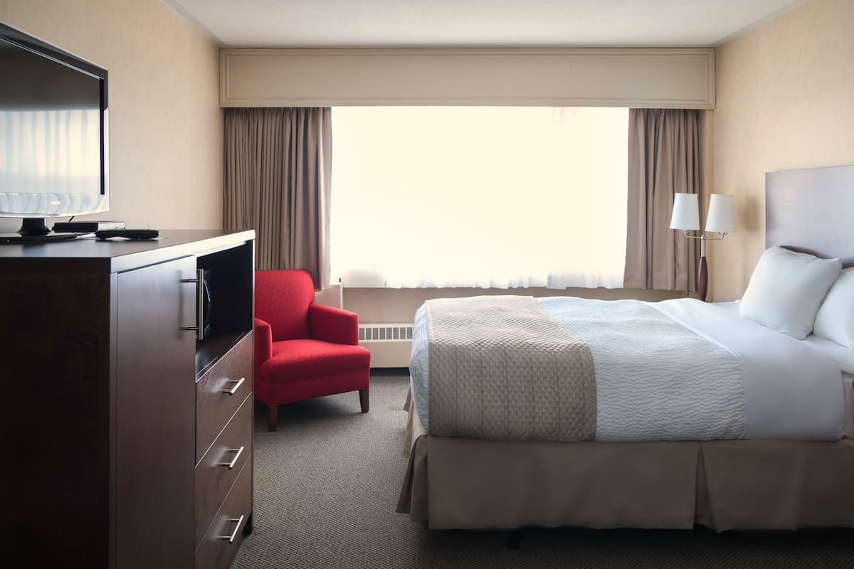Quarto de hotel com cama, televisão, escrivaninha com gavetas, uma cadeira vermelha e duas luminárias.