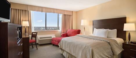 A hotel room with a large bed, a desk, a chair, a TV, and a window with a city view.