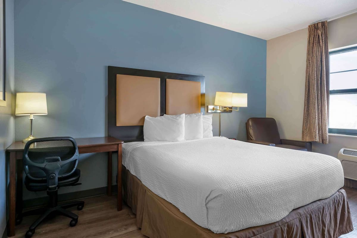 A hotel room with a large bed, a desk with a chair, two wall-mounted lamps, and a window with curtains.