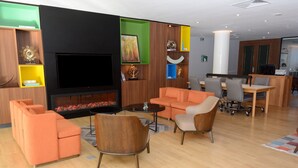 Een moderne hotellobby met een flatscreen televisie, oranje banken, een houten salontafel en een keuken in de achtergrond.